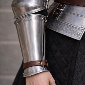 May include: Close-up of polished steel arm armor with decorative etching and a brown leather strap secured with a brass buckle. The armor is part of a medieval costume or historical reenactment ensemble.