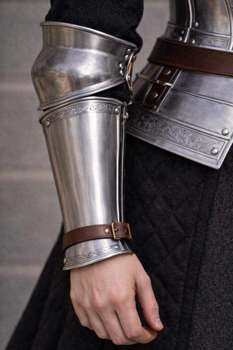 May include: Close-up of polished steel arm armor with decorative etching and a brown leather strap secured with a brass buckle. The armor is part of a medieval costume or historical reenactment ensemble.