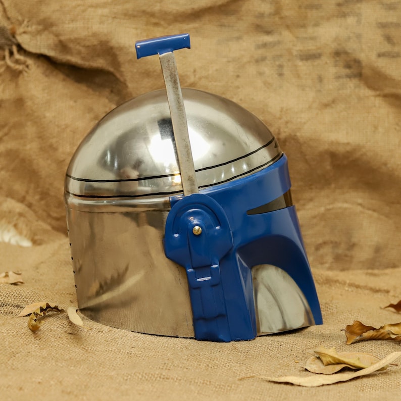 May include: A silver and blue helmet with a T-shaped visor and a blue visor. The helmet has a metallic finish and is likely a costume piece or collectible. The helmet is sitting on a brown surface.