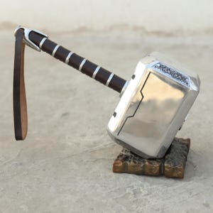 May include: A silver-colored hammer with a black leather strap and a brown wooden base. The hammer has a detailed design on the head and a black leather strap.