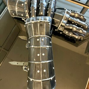 May include: A pair of steel gauntlets, likely for historical reenactment or display. The gauntlets are dark gray with a polished finish, featuring articulated fingers and a segmented cuff. Small rivets secure the plates, and leather straps are visible.
