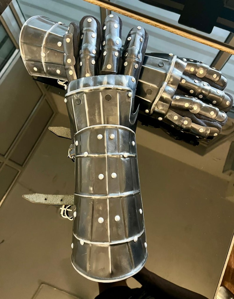 May include: A pair of steel gauntlets, likely for historical reenactment or display. The gauntlets are dark gray with a polished finish, featuring articulated fingers and a segmented cuff. Small rivets secure the plates, and leather straps are visible.