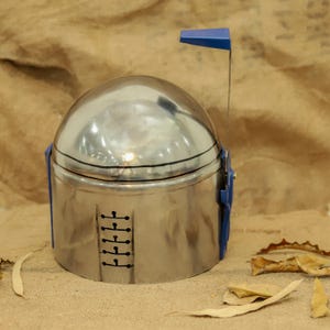 May include: A silver-colored helmet with blue accents and a blue rectangular antenna. The helmet has a rounded top and a rectangular base. Black lines and markings are visible on the helmet's surface. The helmet is likely a costume or prop.