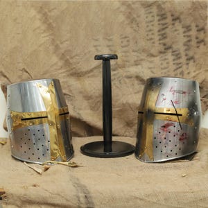 May include: Two silver and gold crusader helmets with a black stand. The helmets have a cross design and are made of metal.