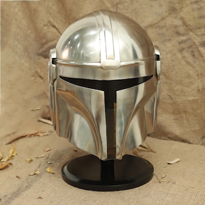 May include: A silver Mandalorian helmet with a black visor, displayed on a black stand. The helmet has a textured surface and a raised ridge on the top.