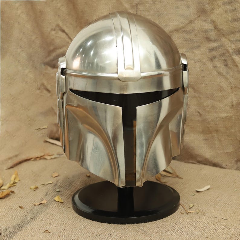 May include: A silver Mandalorian helmet with a black visor, displayed on a black stand. The helmet has a textured surface and a raised ridge on the top.