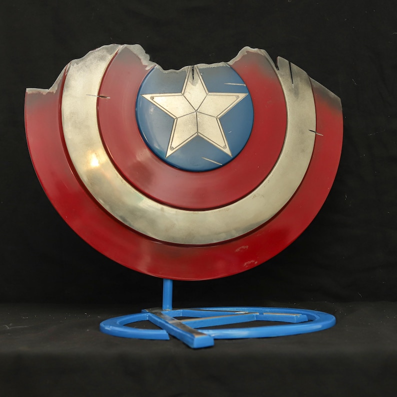 May include: A red, white and blue Captain America shield with a silver star in the centre. The shield is damaged and has a few chips and scratches. It is displayed on a blue stand with a silver A.