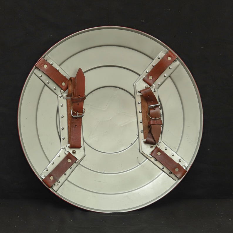 May include: A silver-colored, circular shield with a red border. The shield features concentric circles and a white star in the center. Brown leather straps are riveted to the back for securing the shield.