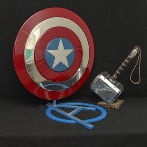 May include: A Captain America shield with red, silver, and blue concentric circles and a white star. Next to it is Thor's hammer, Mjolnir, with a brown handle and a leather strap. A blue Avengers logo is in the foreground.