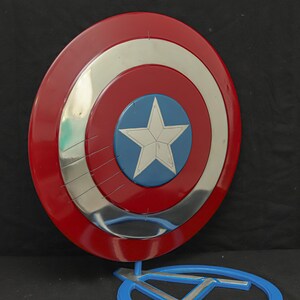 May include: A Captain America shield replica. The shield is round with a red outer ring, a silver middle ring, and a blue center with a white star. The shield is displayed on a blue stand.