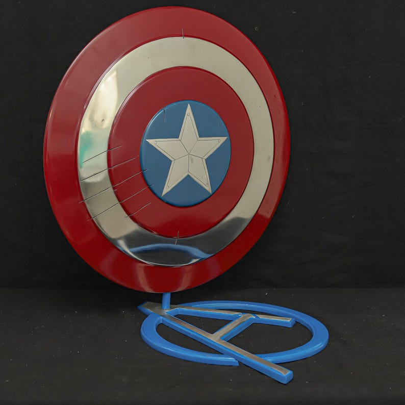 May include: A red, white, and blue shield with a silver band and a blue star in the center. The shield is propped up by a blue stand with a stylized "A" shape. The shield is a replica of Captain America's shield.
