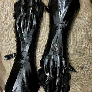 May include: Pair of black, articulated gauntlets with sharp, claw-like extensions. The gauntlets feature intricate detailing and are made of a glossy, dark material. The design suggests a fantasy or cosplay theme.