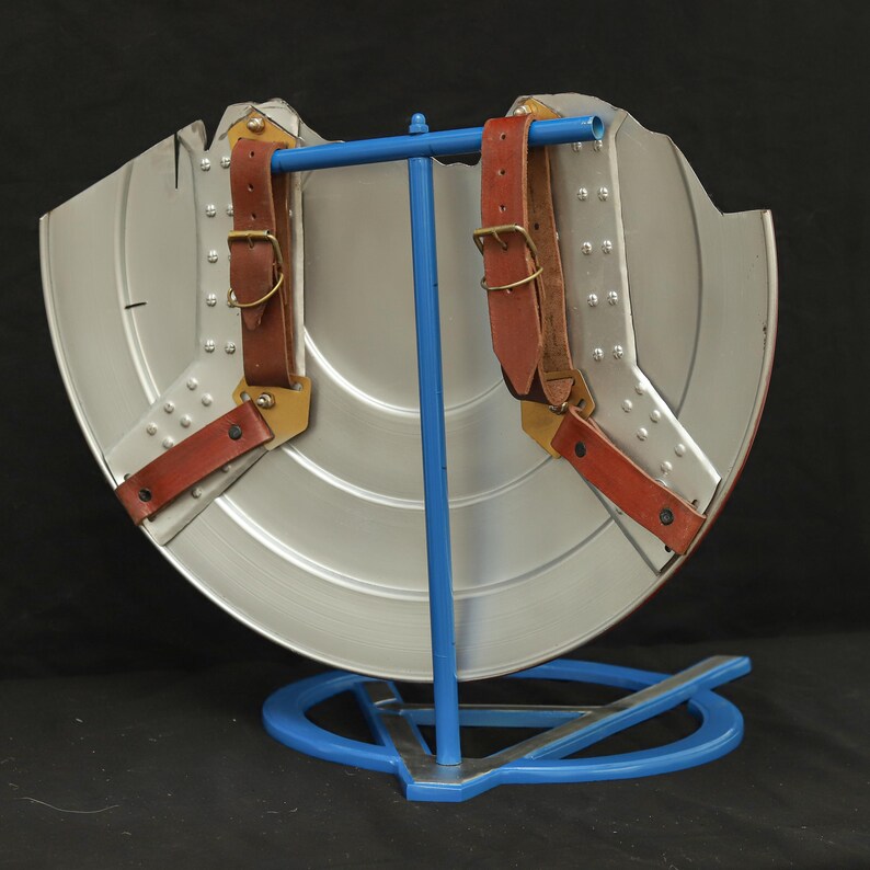 May include: The back of a silver Captain America shield with brown leather straps and gold buckles. The shield is on a blue metal stand.