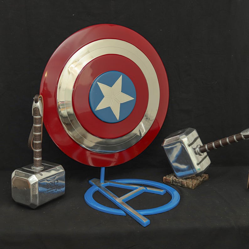 May include: A red, white, and blue Captain America shield with a silver center, a silver hammer with a brown handle, and a blue stand with a silver A symbol.