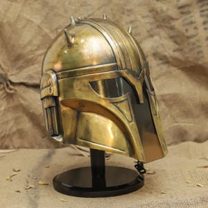 May include: A gold-colored helmet with a black visor and a chain hanging from the bottom. The helmet is on a black stand.