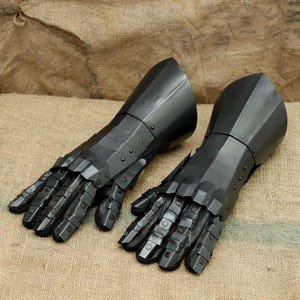 May include: A pair of black metal gauntlets, featuring articulated fingers and a segmented design. The gauntlets are made of dark, reflective metal and are designed to protect the hands and wrists. The background is a neutral tan.