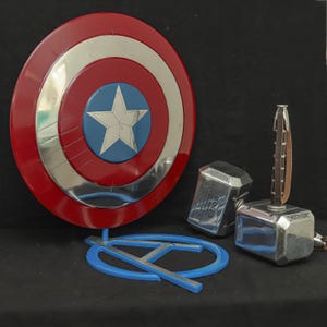 May include: A Captain America shield with a red, silver, and blue design, a blue Avengers logo, and two silver Thor hammers with brown handles. The shield is round with a white star in the center.