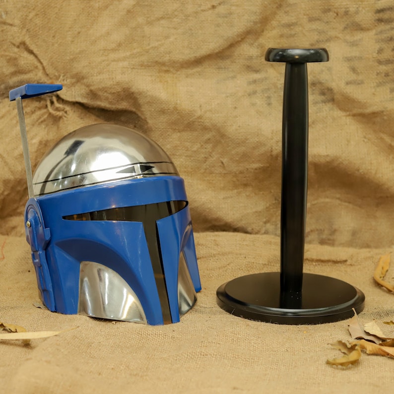 May include: A blue and silver Mandalorian helmet with a black visor and a black helmet stand. The helmet has a silver dome and blue faceplate with black accents. The stand is black with a round base.