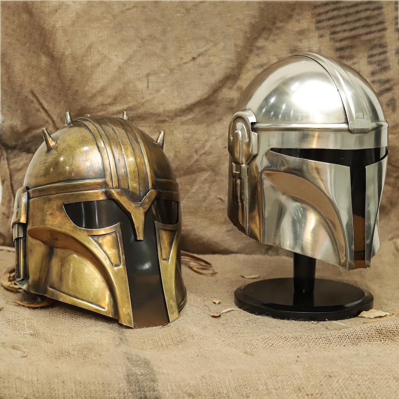 May include: Two Mandalorian helmets, one in a shiny silver finish and the other in a gold finish. Both helmets have a visor and a detailed design.