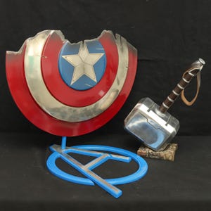 May include: A red, white and blue Captain America shield with a silver metal finish, a silver metal hammer with a brown leather handle, and a blue stand with a silver metal 'A' on it.