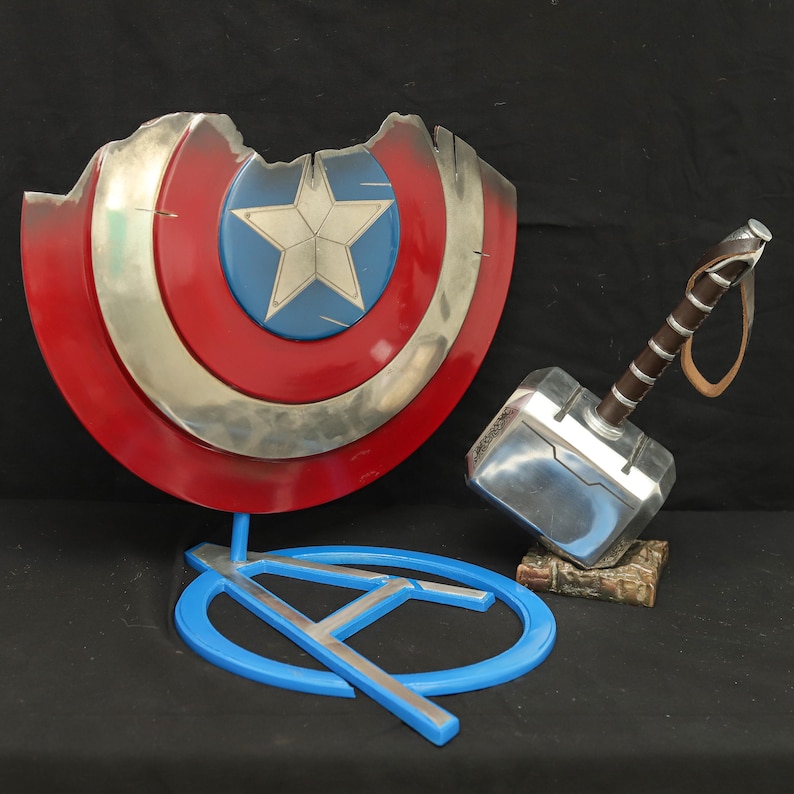 May include: A red, white and blue Captain America shield with a silver metal finish, a silver metal hammer with a brown leather handle, and a blue stand with a silver metal 'A' on it.