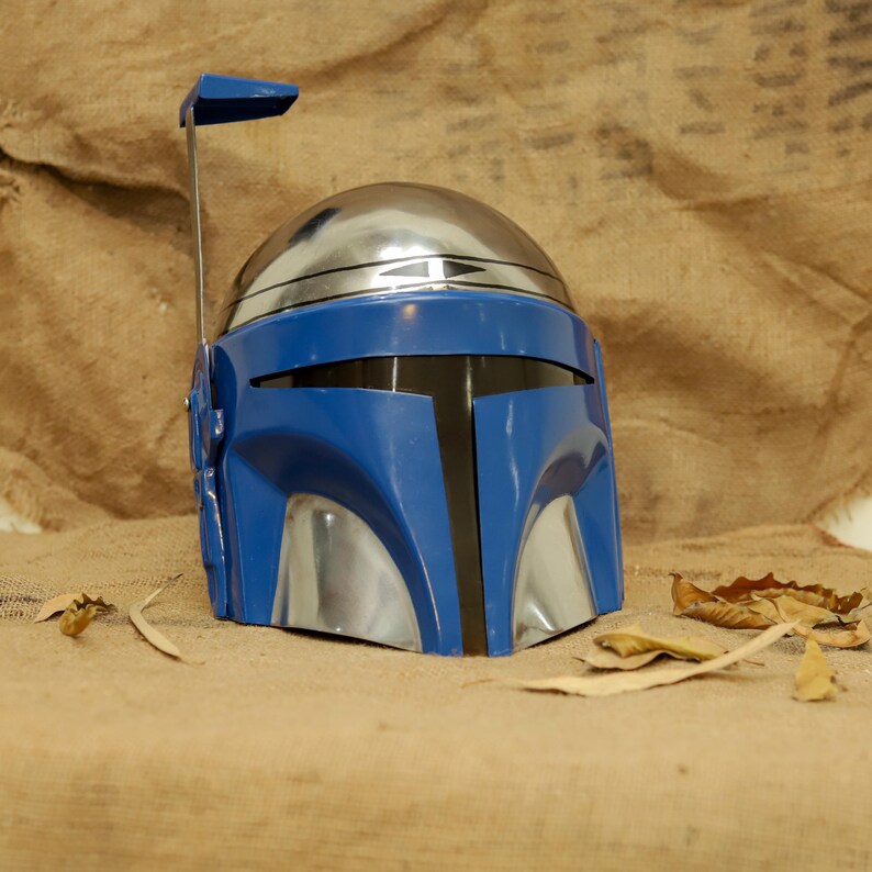 May include: A blue and silver helmet with a visor and a rectangular antenna. The helmet has a shiny, metallic dome and a blue faceplate. The helmet is resting on a tan surface with a few leaves scattered around it.