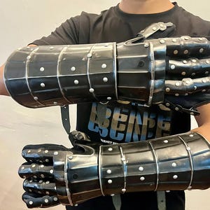 May include: Pair of black metal gauntlets with riveted accents. The gauntlets are segmented, with articulated fingers and wrist protection. They are designed to cover the entire hand and forearm, with leather straps for securing them.