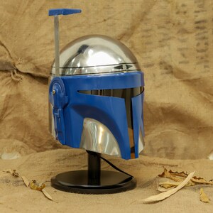 May include: A metallic silver and blue helmet with a T-shaped visor, displayed on a black stand. The helmet has a blue faceplate and a silver dome, with a vertical antenna-like structure extending upwards. The design is reminiscent of a sci-fi character.
