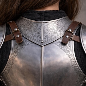 May include: Close-up of a silver chest armor with intricate floral engravings. The armor has brown leather straps and buckles. The design is symmetrical, with a polished finish. The overall theme is medieval or fantasy.