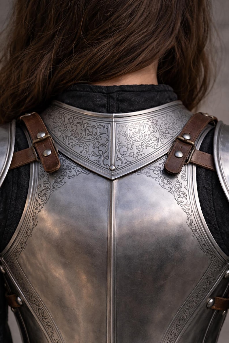 May include: Close-up of a silver chest armor with intricate floral engravings. The armor has brown leather straps and buckles. The design is symmetrical, with a polished finish. The overall theme is medieval or fantasy.