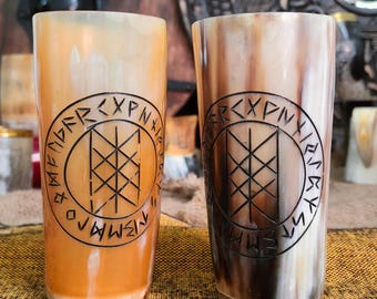 May include: Two tall, cylindrical drinking cups made of brown and white horn with a black design. The design is a circle with runes and a symbol in the center. The cups are on a brown and yellow surface.