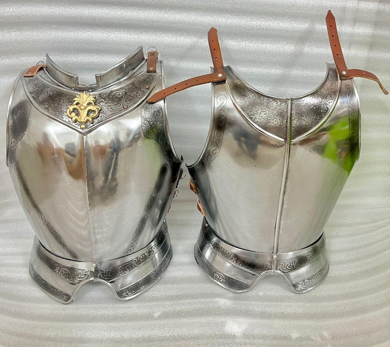 May include: Two polished steel breastplates with intricate engraved designs and brown leather straps. One breastplate features a gold-colored emblem. The armor is designed to protect the torso and is likely for historical reenactment or display.