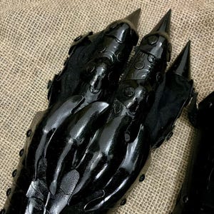 May include: A pair of black, claw-like gloves with sharp, pointed tips. The gloves have a glossy finish and are detailed with textured patterns and rivets. The design evokes a fantasy or cosplay theme.