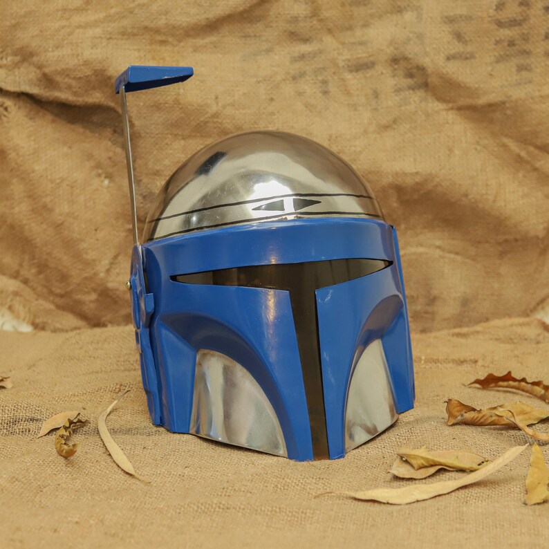 May include: A blue and silver Mandalorian helmet with a T-shaped visor. The helmet has a silver dome and a blue faceplate with silver accents. A blue antenna extends from the top. The helmet is a prop or costume piece.
