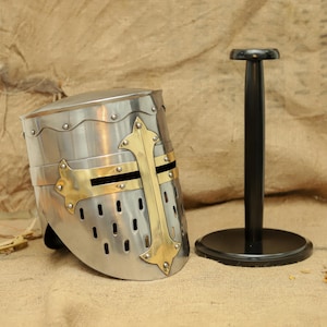 May include: A silver knight helmet with a brass cross design on a black helmet stand. The helmet has a visor and a textured surface.