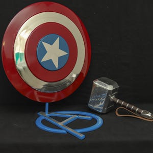 May include: A red, white, and blue Captain America shield with a silver metal center and a blue stand with a silver A. A silver Thor's hammer with a black leather strap is lying on the black surface.