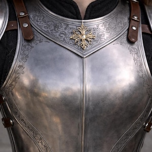 May include: Close-up of a polished steel breastplate with intricate engraved floral patterns and a gold-colored emblem. The armor features brown leather straps and rivets, suggesting a medieval or fantasy theme.