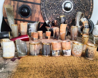 May include: A collection of drinking horns with gold trim and intricate designs. The horns are arranged on a brown surface with a wooden shield in the background.