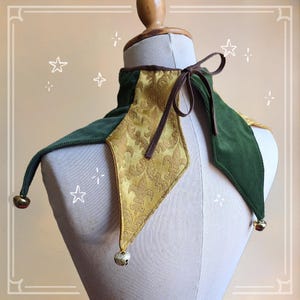 K&ouml;nnte beinhalten: Ein gr&uuml;nes und goldenes Narrenhalsband mit brauner Krawatte. Der Kragen hat ein rautenf&ouml;rmiges goldenes Panel mit floralem Muster, flankiert von gr&uuml;nen Samtabschnitten. Kleine Gl&ouml;ckchen h&auml;ngen an den spitzen Enden und verleihen dem Kost&uuml;maccessoire eine verspielte Note.