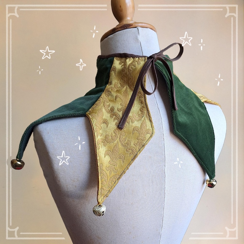 K&ouml;nnte beinhalten: Ein gr&uuml;nes und goldenes Narrenhalsband mit brauner Krawatte. Der Kragen hat ein rautenf&ouml;rmiges goldenes Panel mit floralem Muster, flankiert von gr&uuml;nen Samtabschnitten. Kleine Gl&ouml;ckchen h&auml;ngen an den spitzen Enden und verleihen dem Kost&uuml;maccessoire eine verspielte Note.
