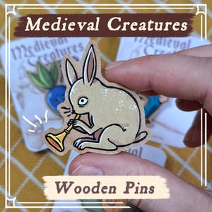 Puede incluir: Pin de madera con un conejo de dibujos animados tocando la trompeta. El conejo es de color marrón claro con manchas oscuras. El pin está etiquetado como "Medieval Creatures" y "Wooden Pins".