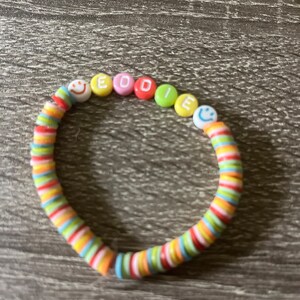 Peut inclure: Un bracelet de perles colorées avec le nom "EDDIE" épelé avec des perles de lettres. Le bracelet présente un mélange de perles en forme de disque aux couleurs de l'arc-en-ciel et de perles smiley. Le bracelet est posé sur une surface en bois.