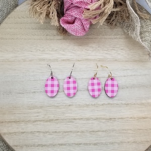 May include: Oval-shaped earrings with a pink and white gingham pattern. The earrings are displayed on a light wooden surface, with a pink fabric and burlap ribbon in the background. The earrings have silver and gold hooks.