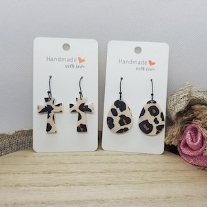 May include: Two pairs of dangle earrings with leopard print designs. One pair features a cross shape and the other pair features a teardrop shape. The earrings are handmade with love.