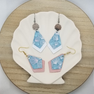May include: A pair of dangle earrings with a blue and white seashell pattern. The earrings are made of leather and have a pink backing. The earrings are displayed on a wooden plate.