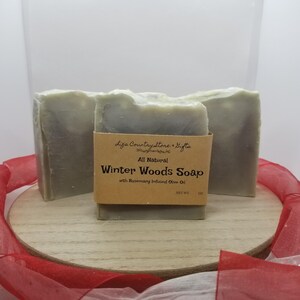 May include: Three bars of handmade soap with a light green hue, presented on a wooden surface. The central bar features a brown label that reads "Winter Woods Soap" with "Rosemary Infused Olive Oil". The soap is a natural product.