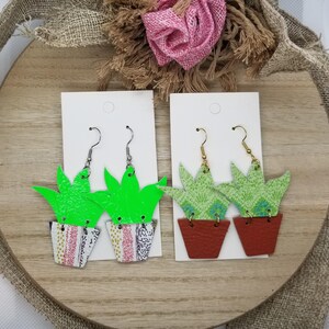 May include: Two pairs of dangle earrings featuring a green succulent plant in a patterned pot. One pair has a bright green succulent and the other has a light green succulent.