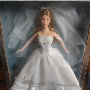 Peut inclure: Une poupée de mariée blanche portant une robe de mariée blanche avec un voile et tenant un bouquet de fleurs blanches. La poupée est dans une boîte avec le texte "Millennium Wedding The Bridal Collection" sur le devant.
