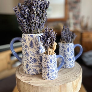May include: Three ceramic jugs with blue handles and a white and blue floral and rabbit pattern, each filled with dried lavender. The jugs are arranged on a wooden stool, creating a rustic and charming display.