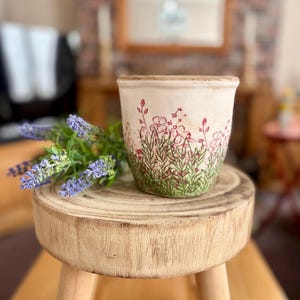 May include: A cream-coloured ceramic flower pot with a floral design featuring pink flowers and green grass. The pot sits on a small wooden stool, with a sprig of lavender to the side. The pot is a decorative item.